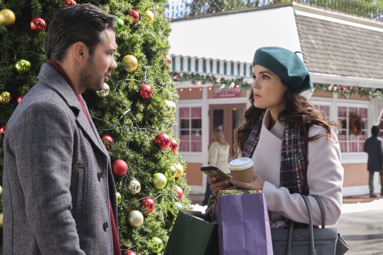 Still from A Timeless Christmas depicting a man (left) with dark hair, wearing a light gray coat, looking over at a woman (right) wearing a green beret, dark gray scarf, and a white sweater, holding onto multiple paper shopping bags and a coffee cup, standing in front of a decorated tree with string lights and red and gold ornaments.