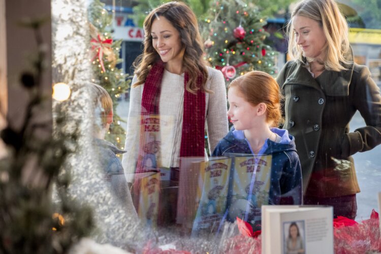 Still from A Bramble House Christmas depicting a woman (left) with brown curly hair, wearing a white sweater and a red scarf, another woman (right) with blonde hair, wearing a gray button-up coat, and a young girl (middle) with ginger hair and a blue zip-up jacket in front of a glass store window.
