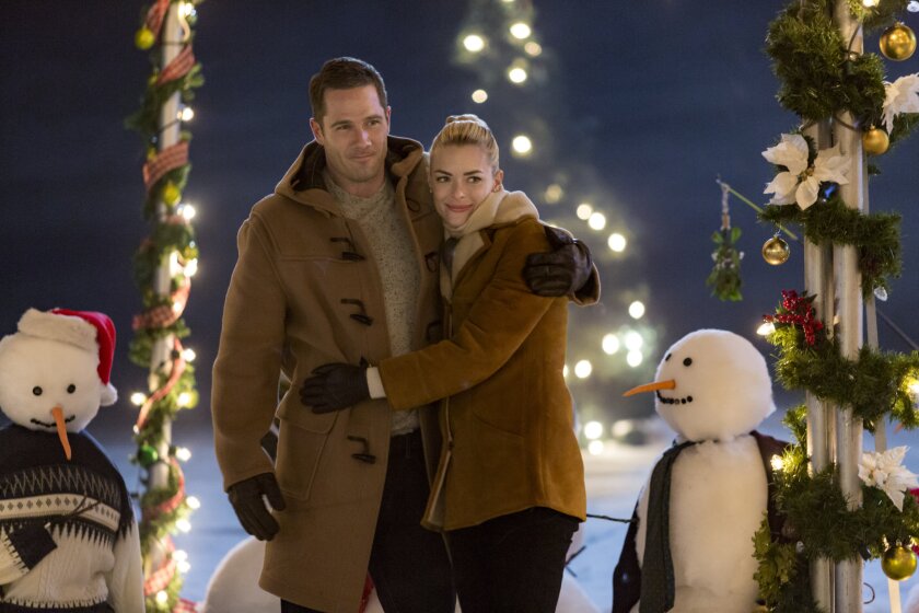 Still from The Mistletoe Promise depicting a man (left) with a light brown coat hugging a woman (right) with blonde hair and a brown jacket. On the far left is a snowman wearing a blue and gray decorative sweater and a red Santa hat, while on the far right is a snowman wearing a black cardigan.