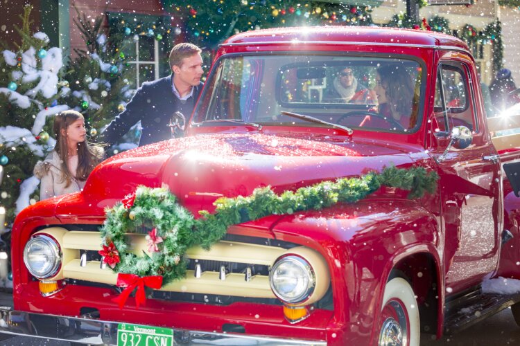 Still from Christmas in Evergreen depicting a woman inside a red truck, decorated with a green garland and a green wreath with a red bow. On the left is a man in a dark blue coat and a young girl wearing a white coat, looking into the window of the truck.