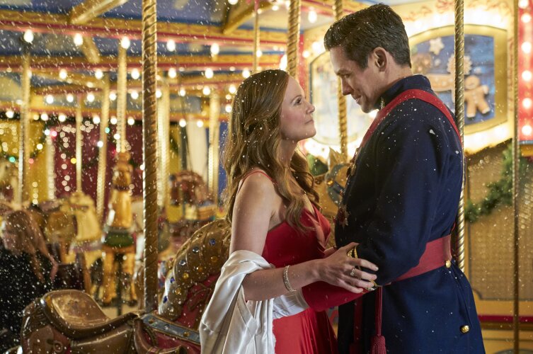 Still from A Christmas Carousel depicting a woman (left) wearing a sleeveless red dress and a white shawl, looking up and holding onto a man (right) wearing a blue coat with red adornments, standing in front of a lit-up carousel.