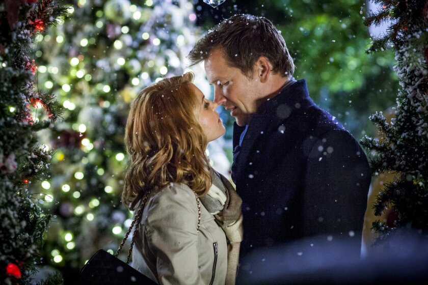 Still from A Christmas Detour depicting a woman (left) with ginger hair wearing a gray coat, leaning up to kiss a man (right) with short brown hair, wearing a black coat. The border of the image is snow-covered trees with red string lights on them.